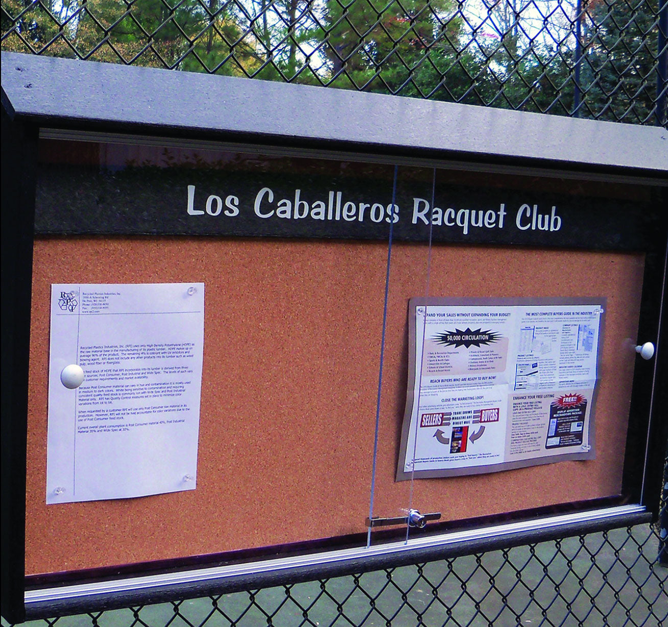 Outdoor community bulletin board with cork surface, sliding lockable acrylic doors, mounted on chain-link fence