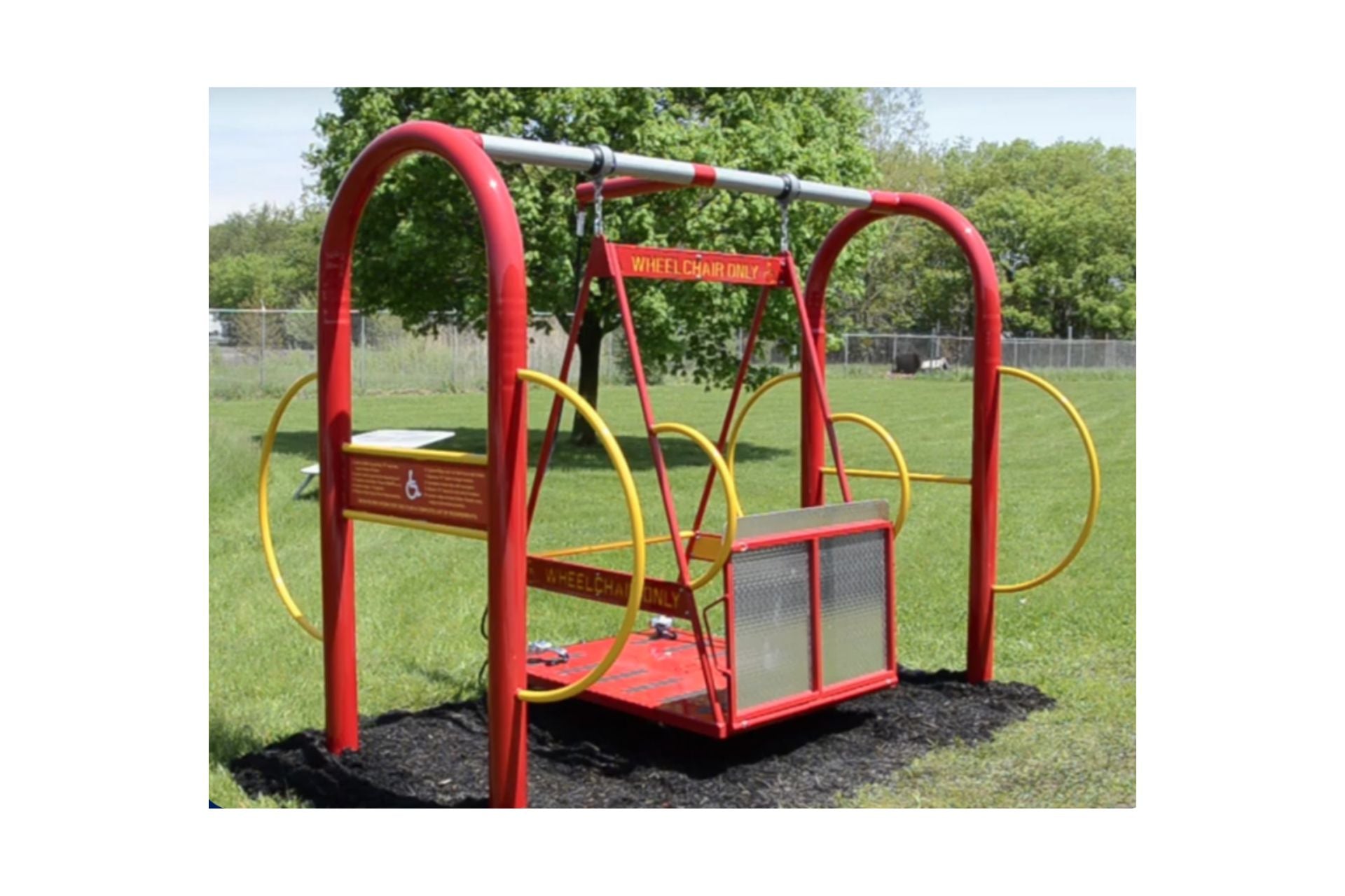 Red and yellow wheelchair-accessible swing set platform with safety rails, locking mechanisms, and grass surface