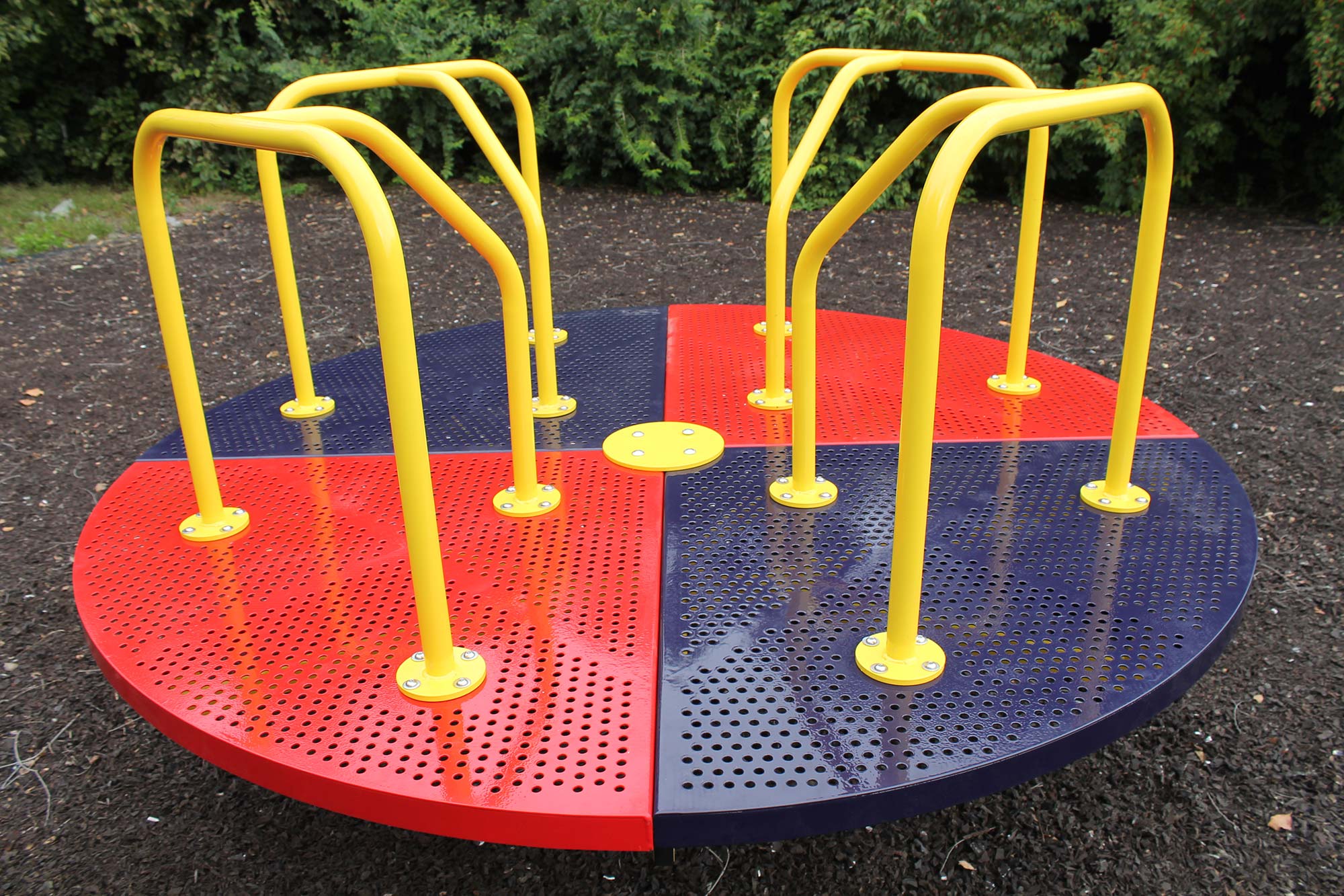 Colorful playground merry go round with red and navy perforated steel deck and yellow handrails on mulch surface