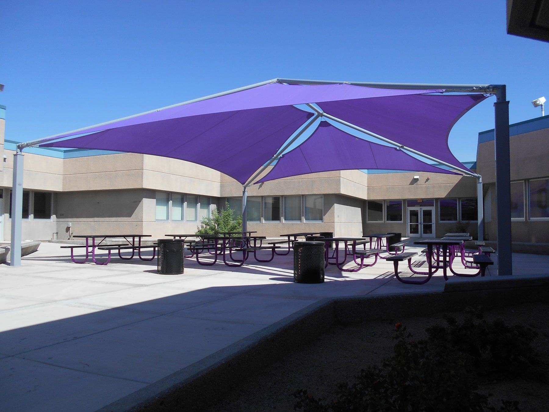 SuperSpan Hip Multi-Panel Shade Structure with vibrant purple fabric panels over purple picnic tables and trash bins in a school courtyard