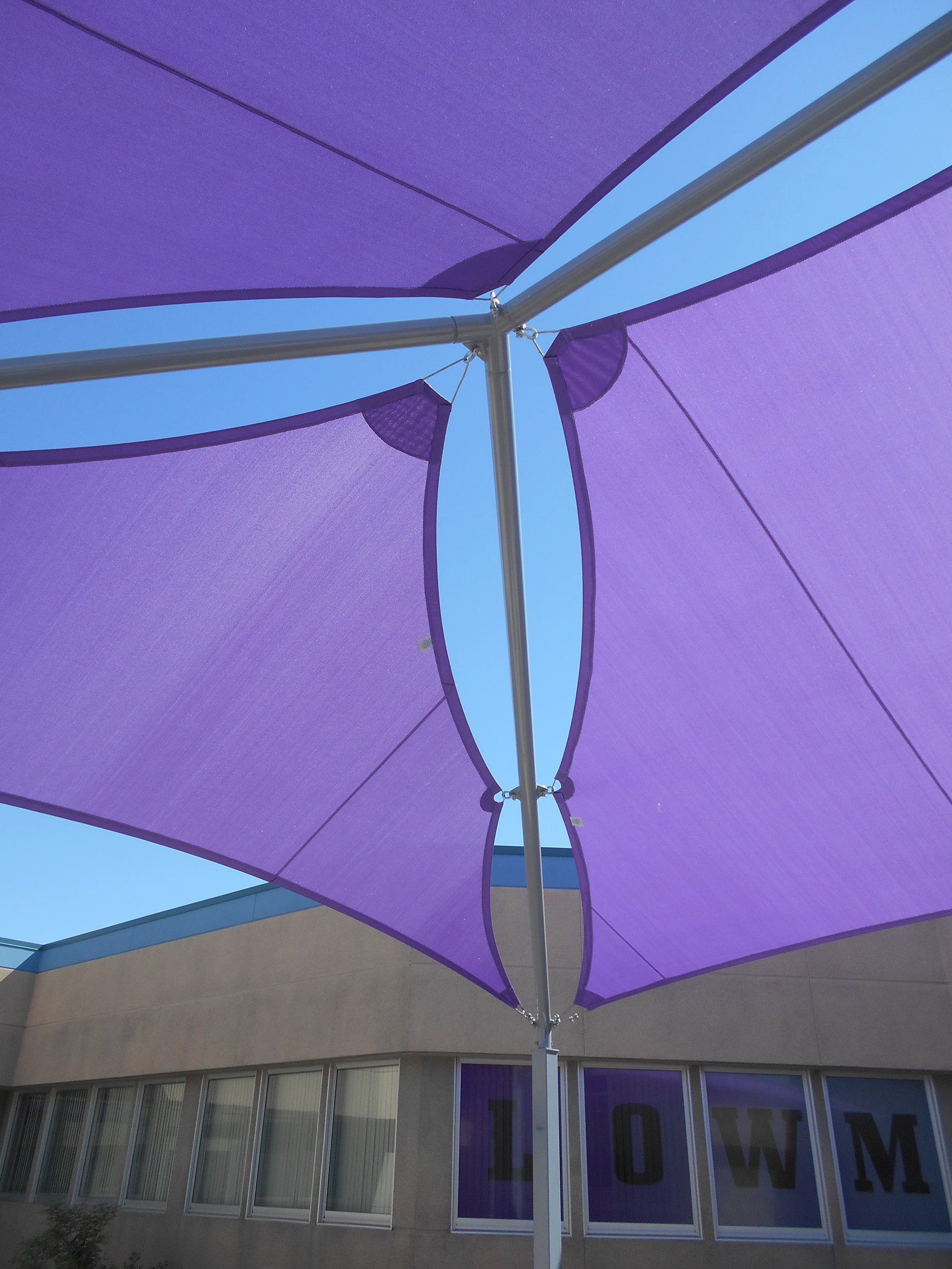 Purple multi-panel fabric shade structure with silver metal posts over a school building exterior