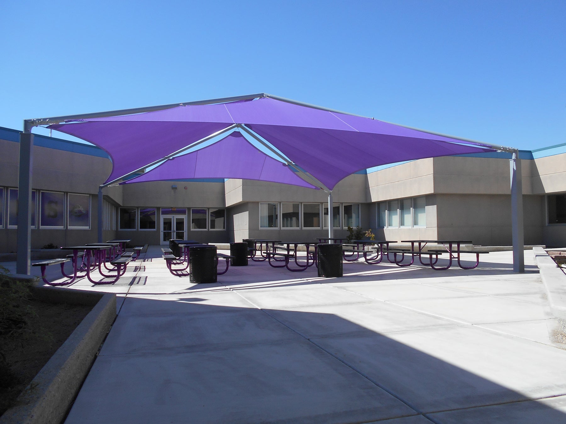 Large four-post shade structure with vibrant purple multi-panel fabric over purple picnic tables in a school courtyard