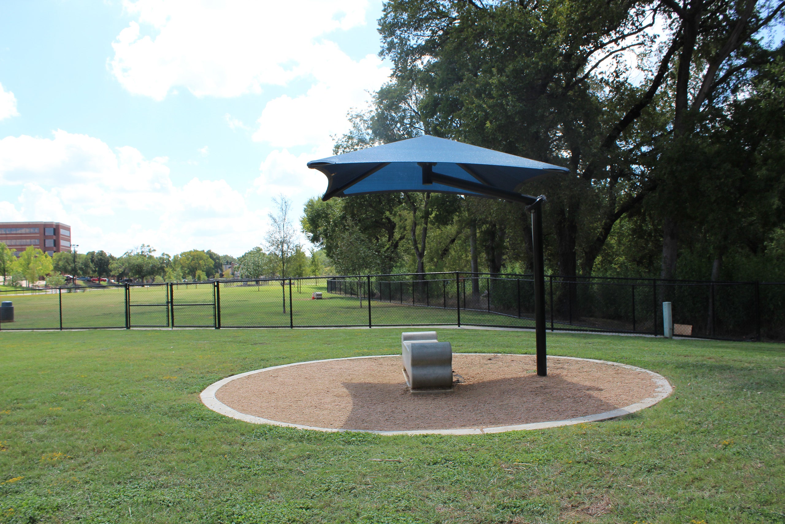 Single post cantilever shade structure with blue pyramid canopy over circular sand area and bench in park
