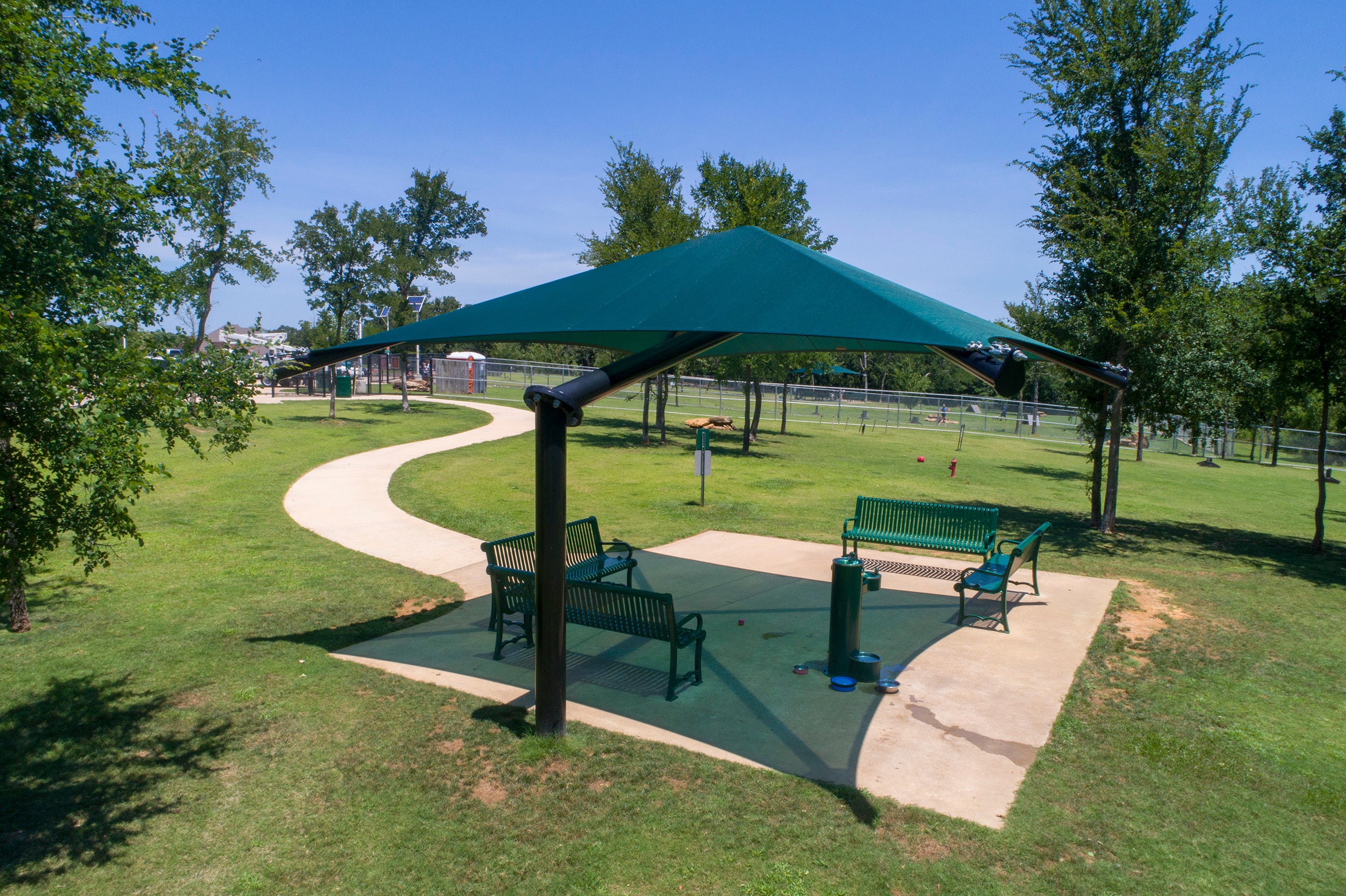 Single post pyramid cantilever shade with green fabric canopy over park benches on concrete pad in grassy area