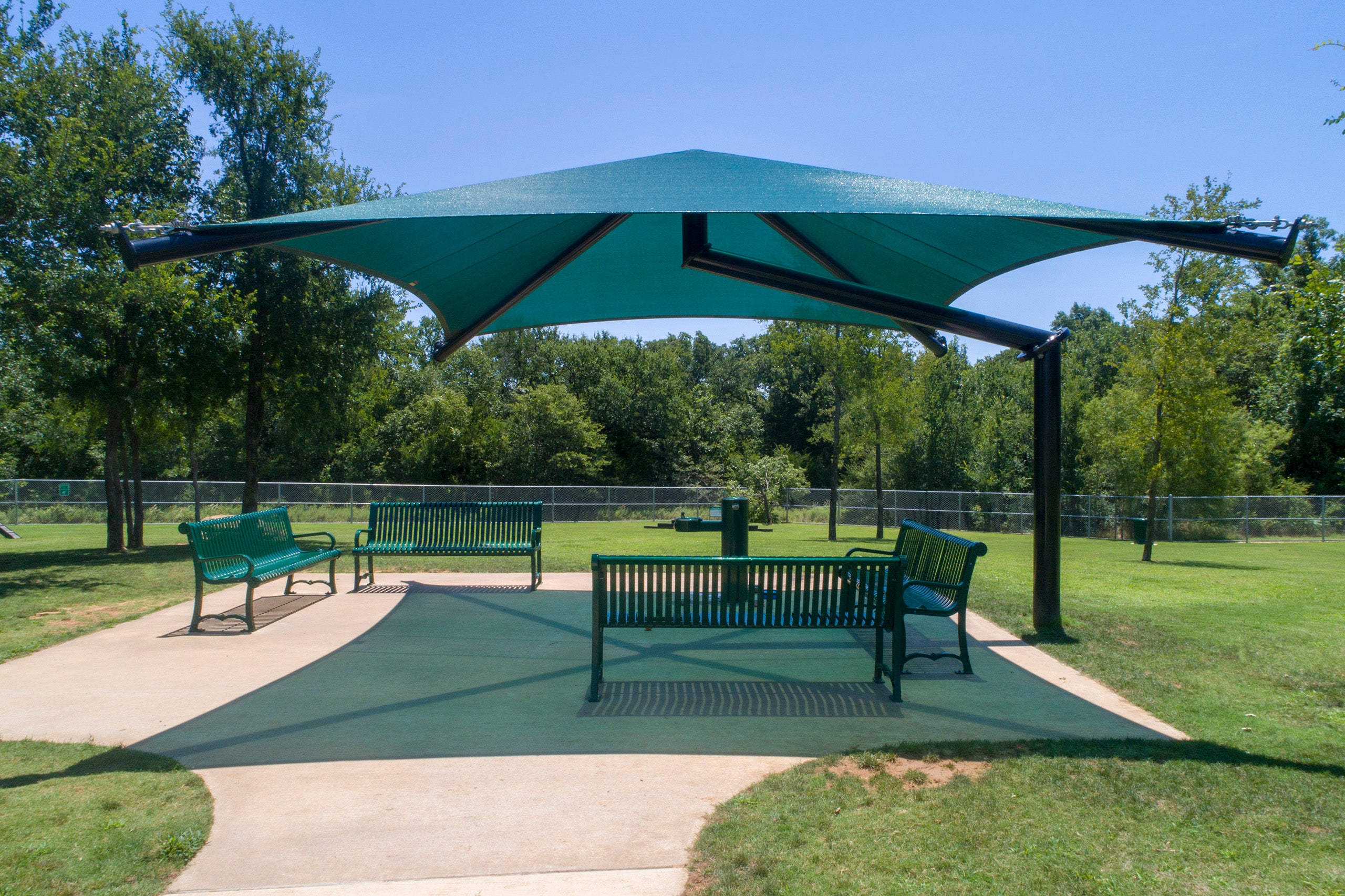 Single Post Pyramid Cantilever Shade Structure with teal UV-blocking fabric over green park benches on concrete pad in sunny park