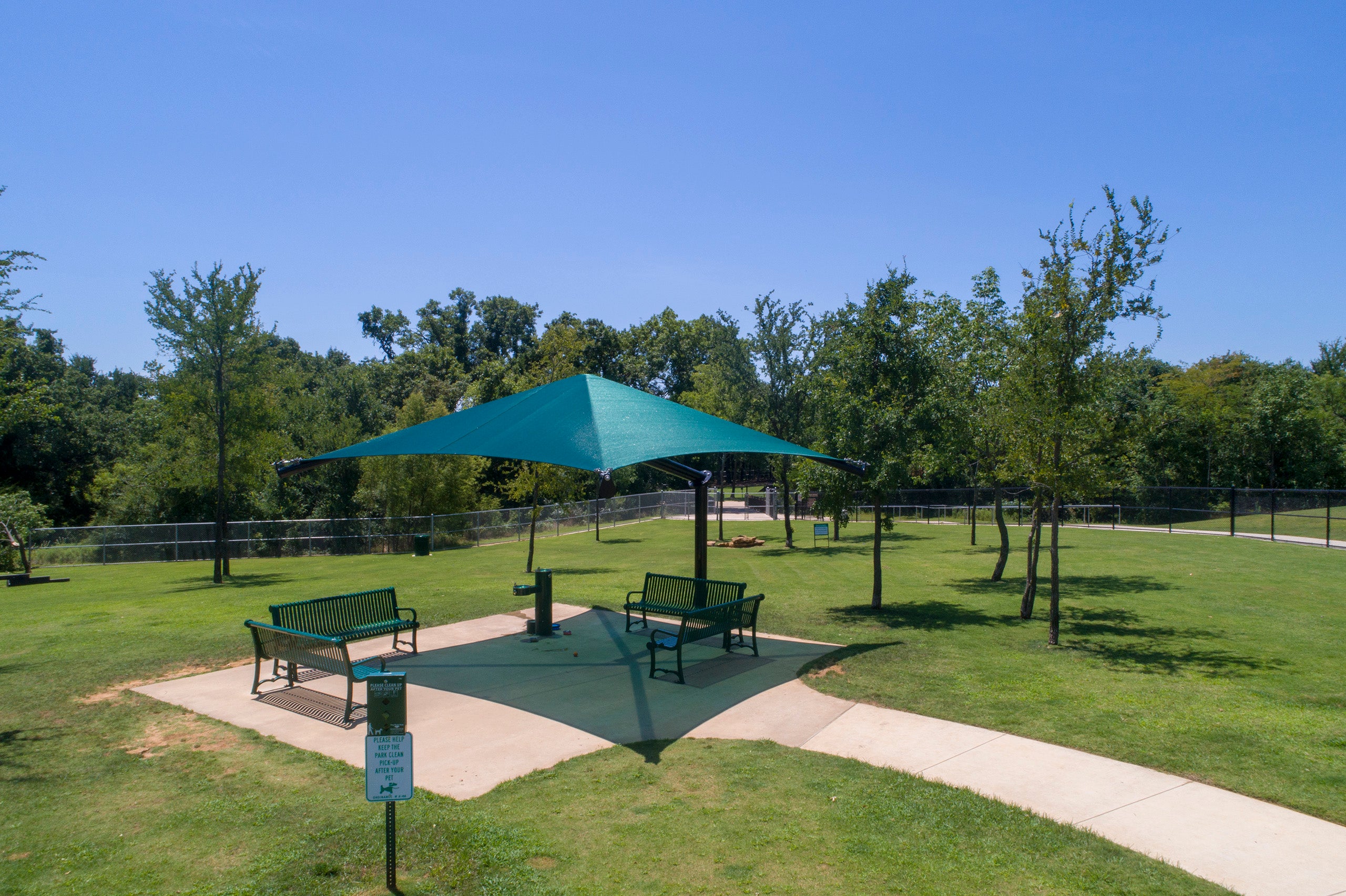 Single post cantilever shade structure with teal pyramid canopy over benches in a green park setting