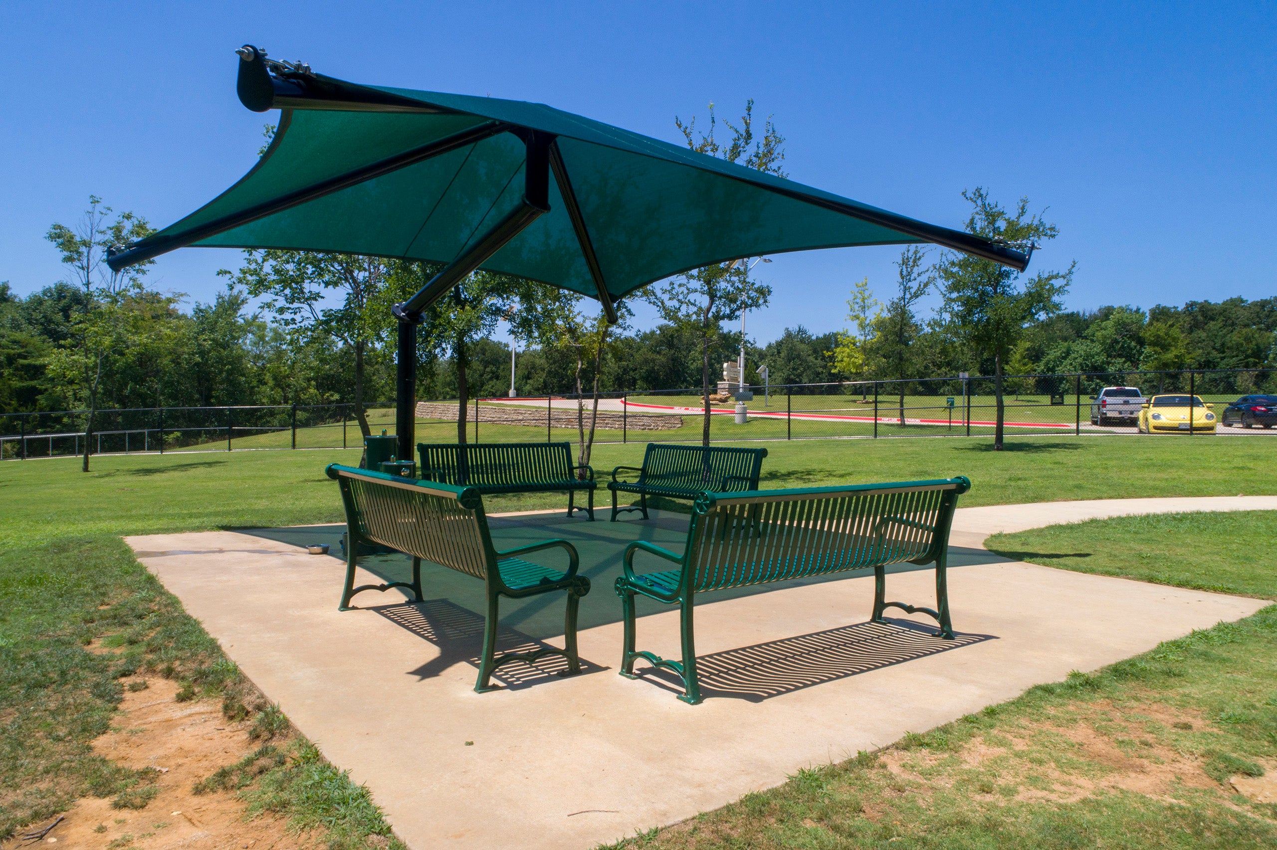 Single post cantilever shade structure with green pyramid fabric canopy over four green metal benches on concrete pad in park setting
