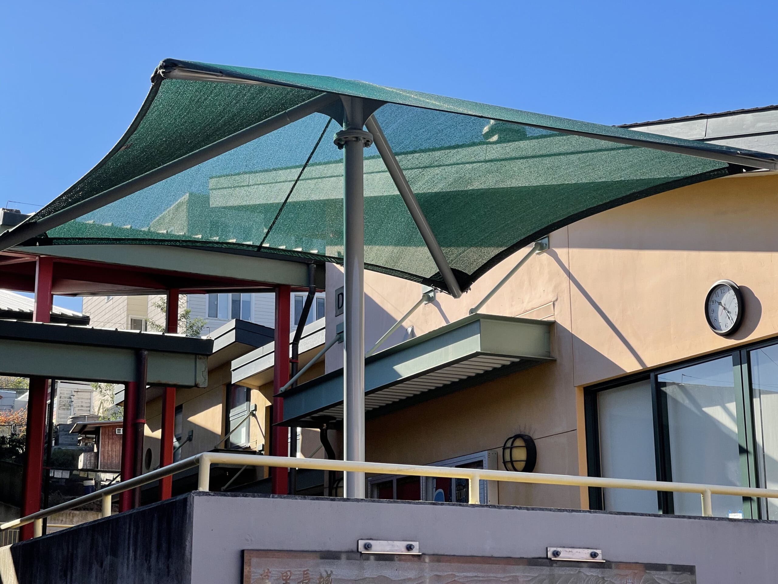 Green single-post pyramid shade structure with UV-blocking fabric over outdoor play area near building