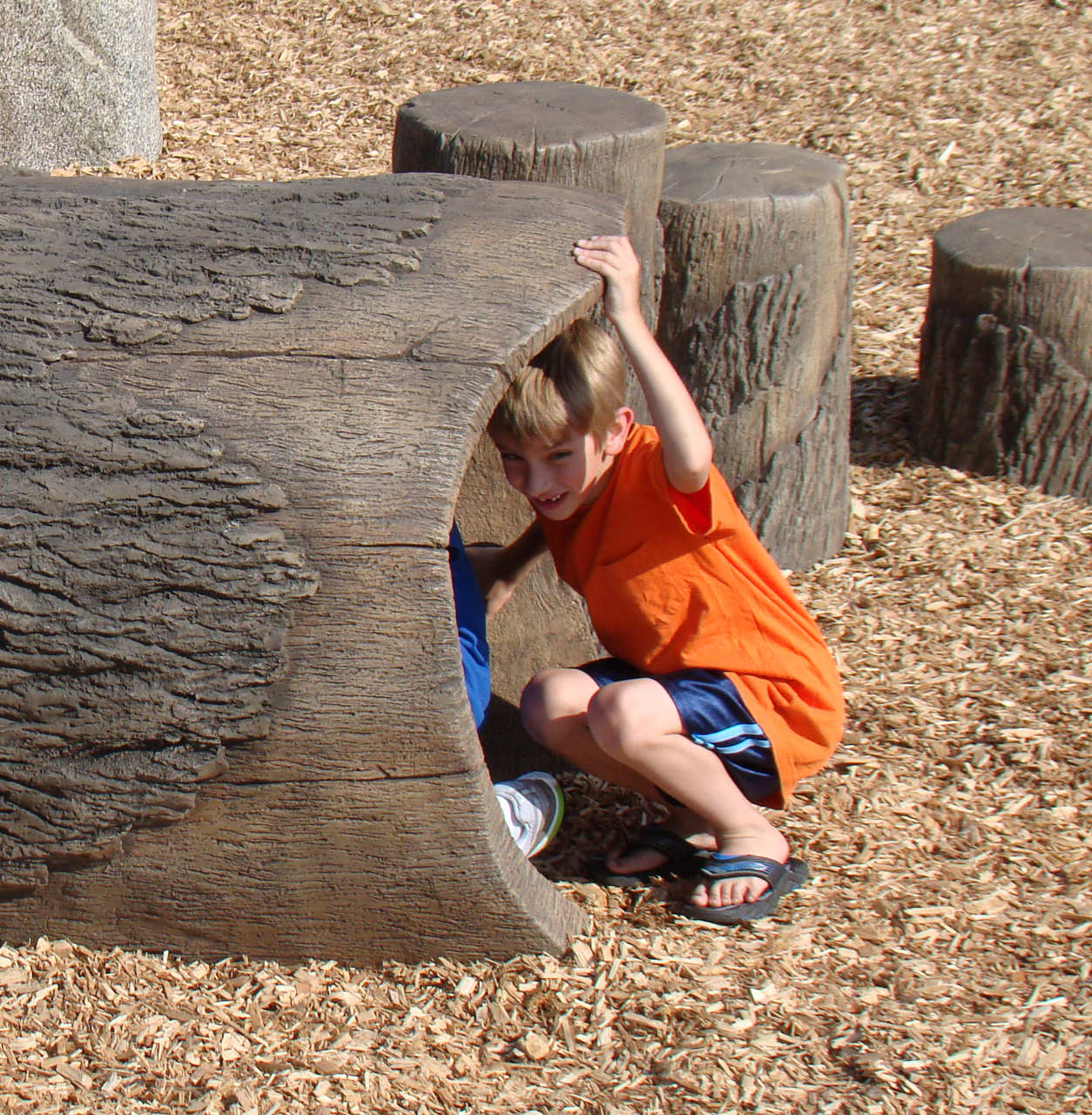 Child inside a realistic wood grain playground crawl tunnel with tree stump climbing elements on wood chip ground