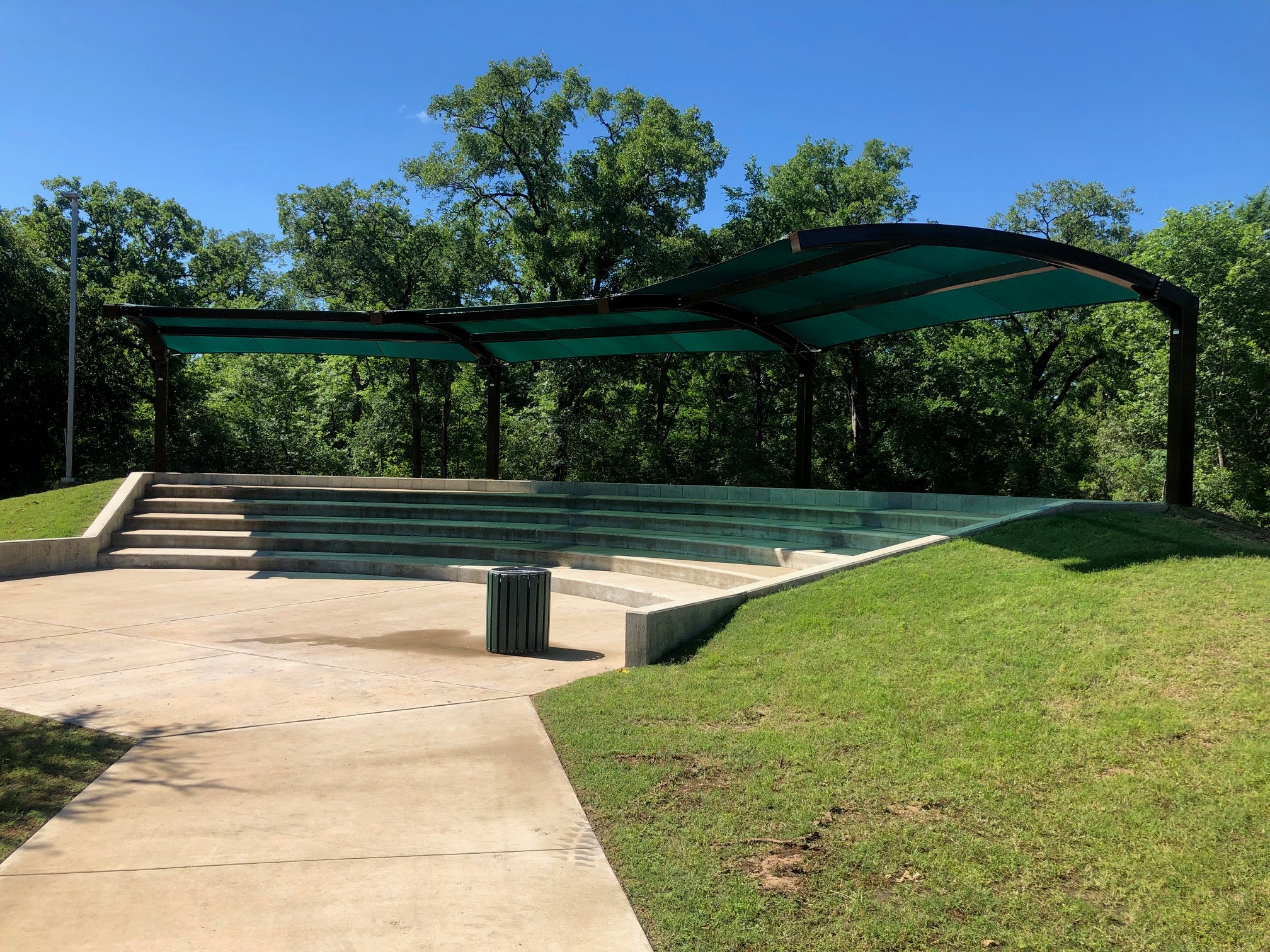 Cantilever shade structure with arched roof over concrete amphitheater seating surrounded by trees and grass