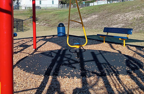 Black circular playground wear mat beneath yellow swing frame surrounded by wood chips and playground equipment
