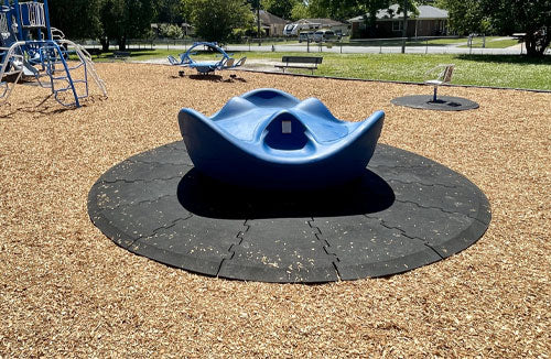 Playground spinner with blue molded seat on circular black rubber wear mat surrounded by wood chips