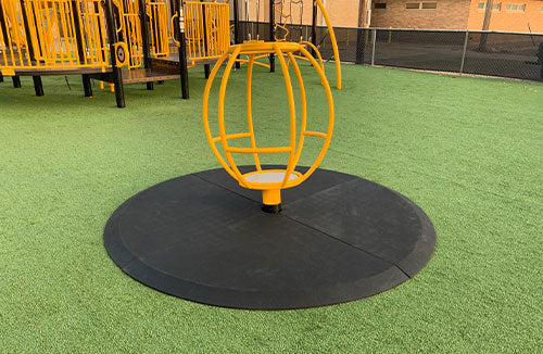 Black multi-piece protective mat with beveled edges beneath a yellow spherical playground spinner on green safety surfacing
