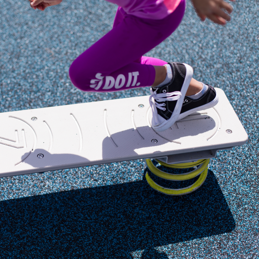 Child in purple leggings balancing on white spring-mounted board with textured grip on blue playground surface