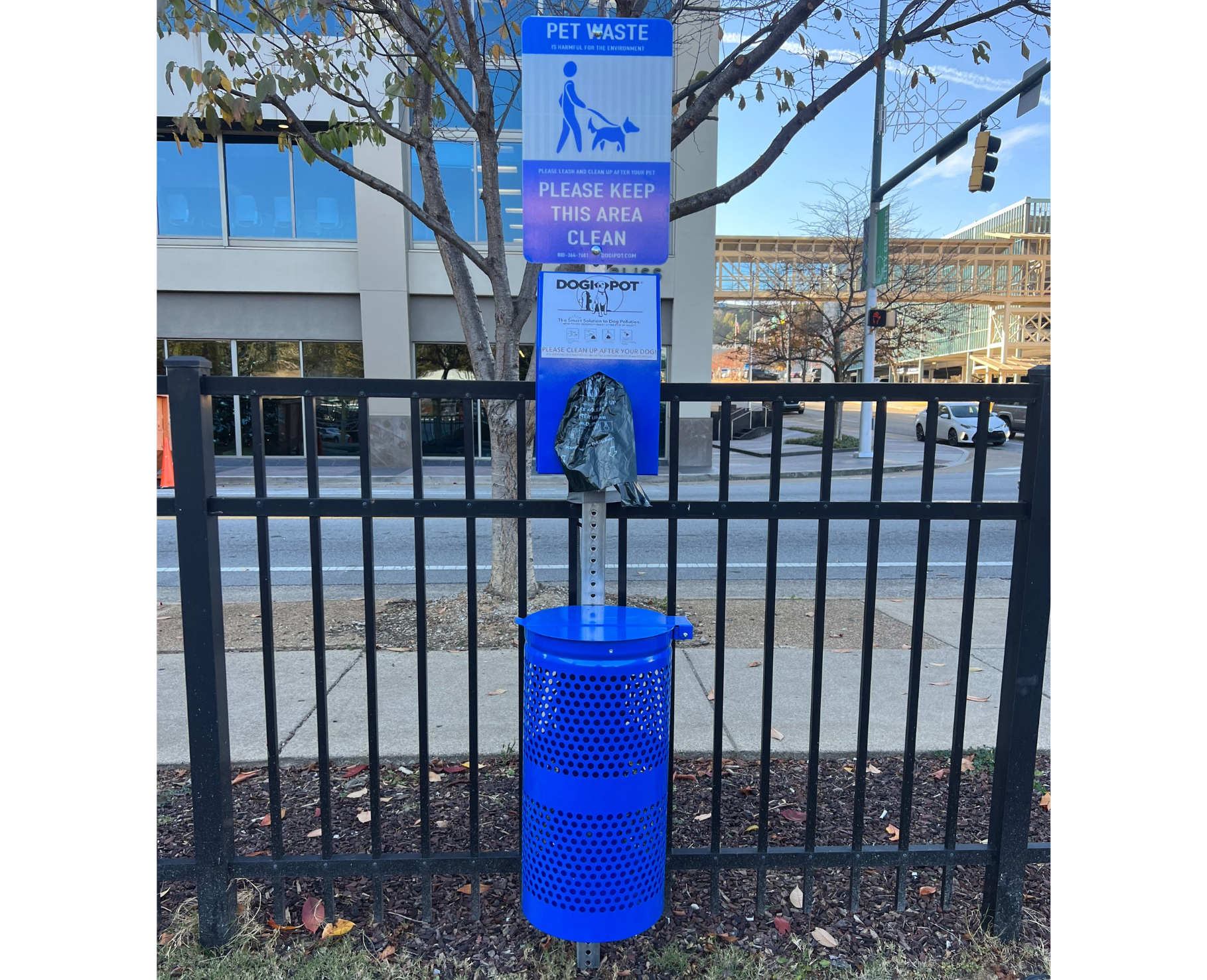 Blue aluminum pet waste station with perforated receptacle, leash-and-dog icon, and bag dispenser mounted on black fence