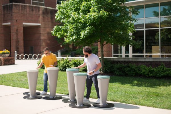 Two men playing outdoor powder-coated steel percussion drums with synthetic drumheads on a paved area near greenery