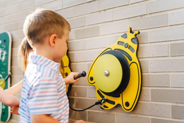 Child playing yellow bee-shaped outdoor musical instrument with mallet, mounted on wall, engaging in interactive music play