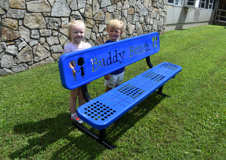 Blue playground bench with perforated game patterns and child figure cutouts, two children standing behind on grass