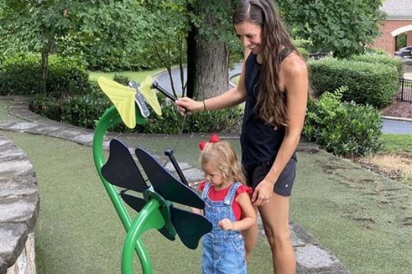 Toddler playing colorful butterfly-shaped outdoor musical instrument with adult assistance on green playground surface