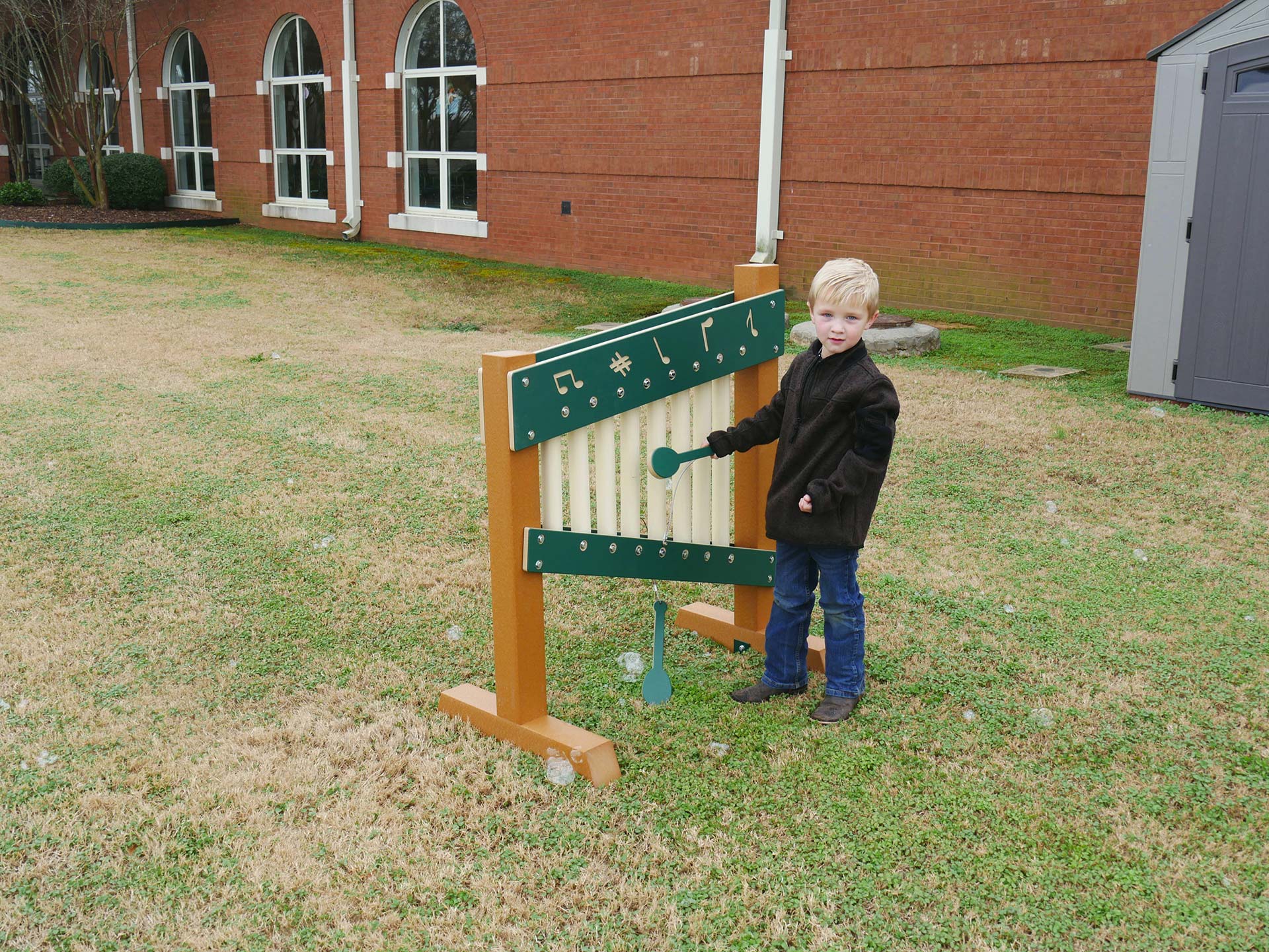 Child playing colorful freestanding outdoor musical chime wall with mallets on grass near brick building