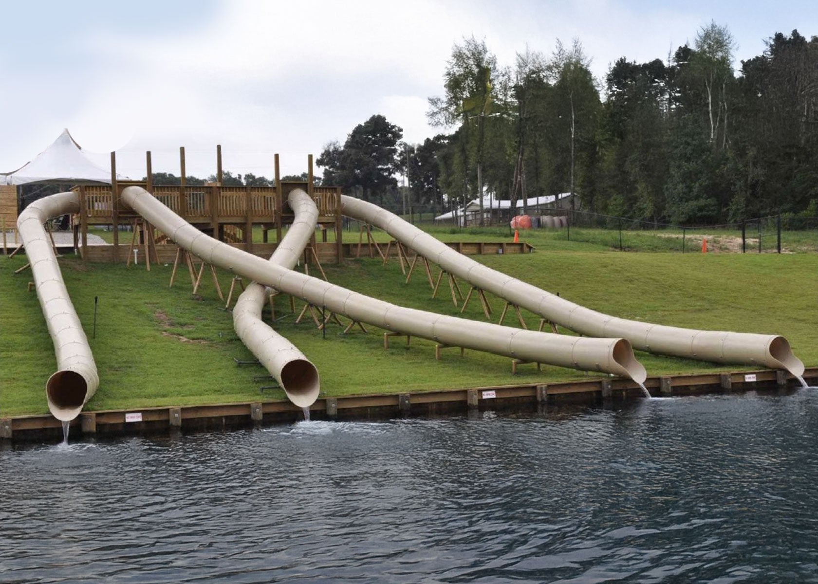 Four beige commercial water slides descending a grassy hill into a lake, supported by wooden framework and deck platform