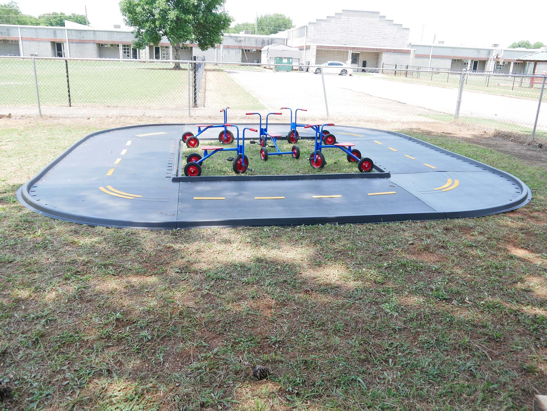Outdoor ADA-compliant trike path with black surface, yellow markings, and six colorful tricycles on grass