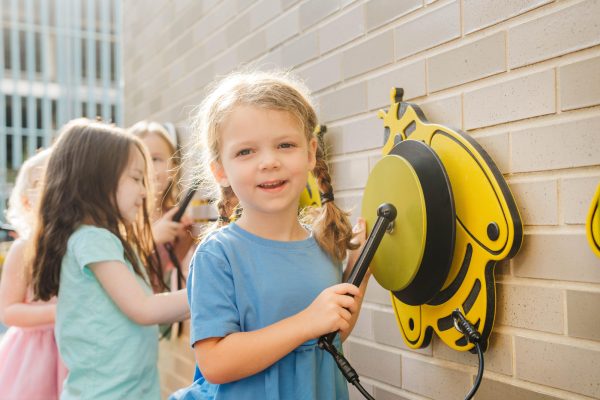 Young girl in blue shirt playing bee-shaped percussion on wall-mounted honeycomb metallophone in outdoor play area with children nearby