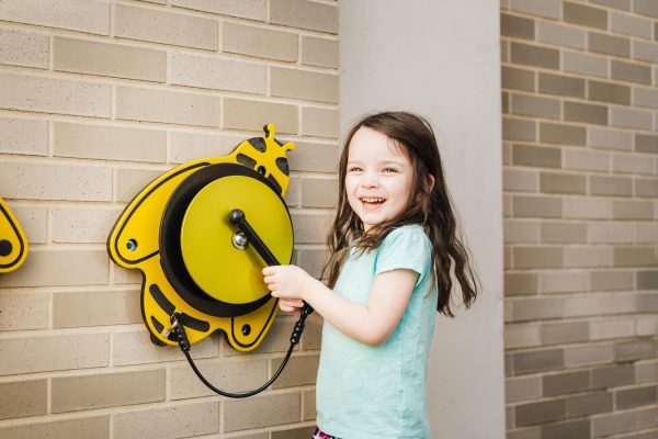 Smiling girl playing yellow bee-shaped outdoor musical instrument mounted on brick wall, bright and engaging playground feature