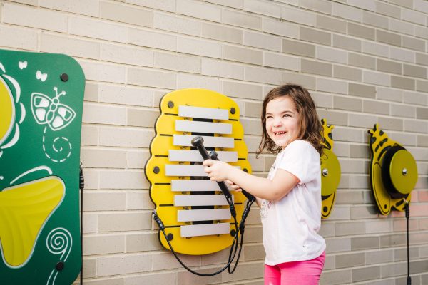 Young girl playing yellow honeycomb metallophone with bee-shaped percussion on outdoor brick wall playground equipment