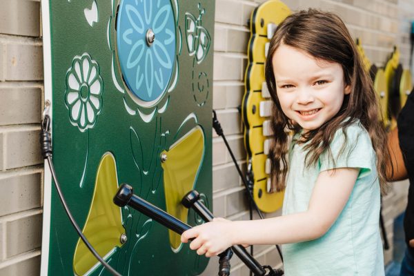 Child with brown hair and blue shirt playing an outdoor green musical panel with botanical designs and tethered mallets mounted on a brick wall