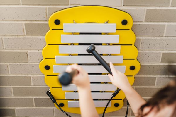 Beehive-shaped outdoor metallophone with white bars and tethered mallets mounted on brick wall, child’s hands playing