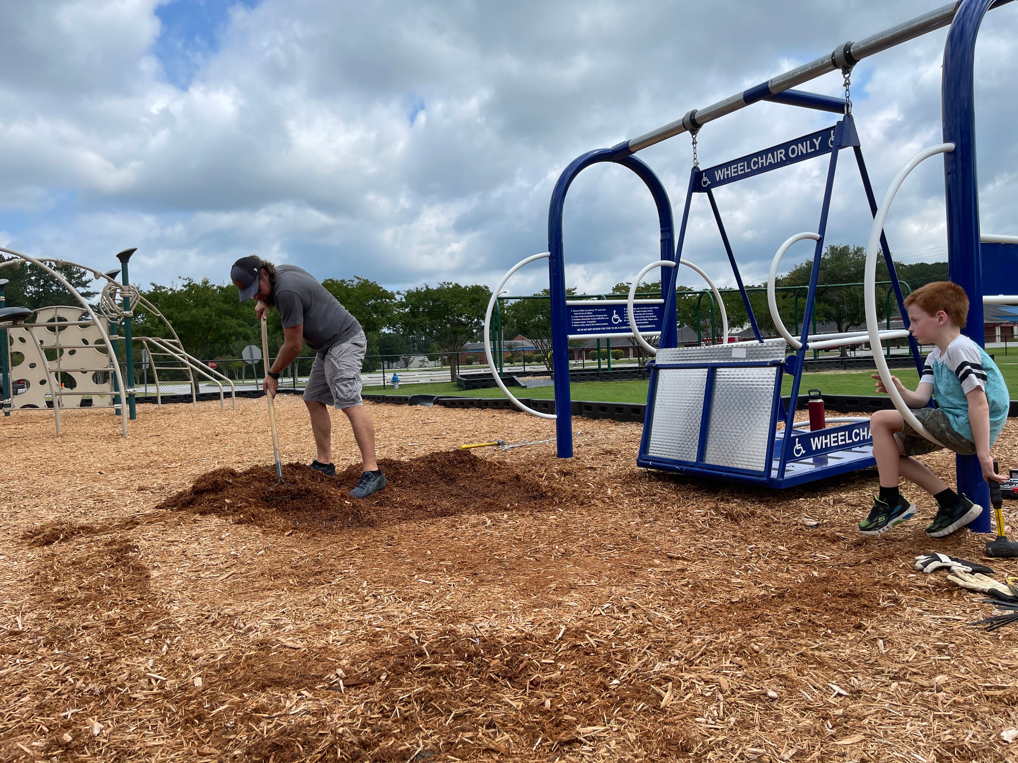 Blue wheelchair-accessible swing platform with safety rings on playground wood chips, child nearby, man raking mulch