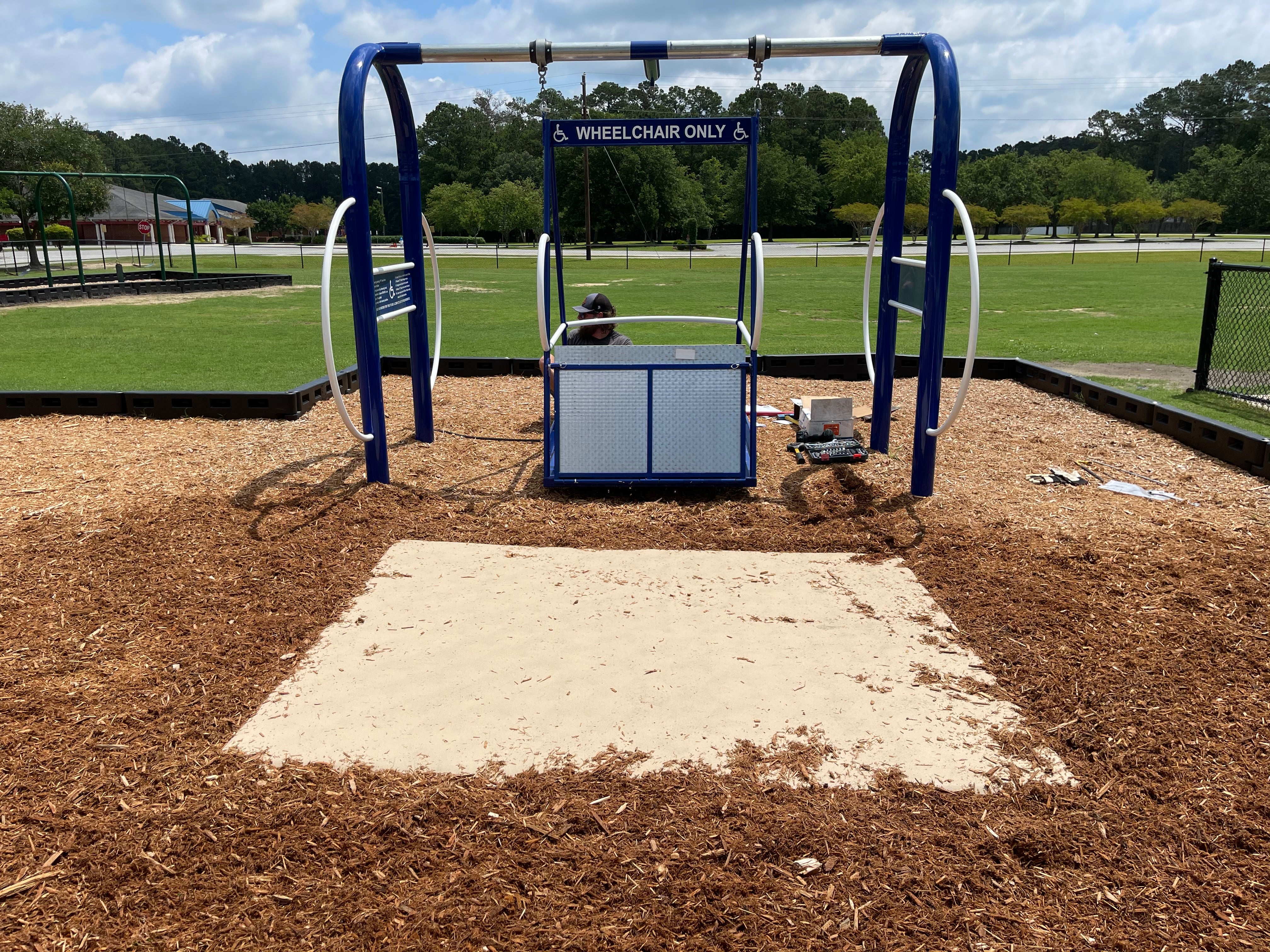 Blue wheelchair-accessible commercial swing set platform with safety rails and ground mat in outdoor park setting
