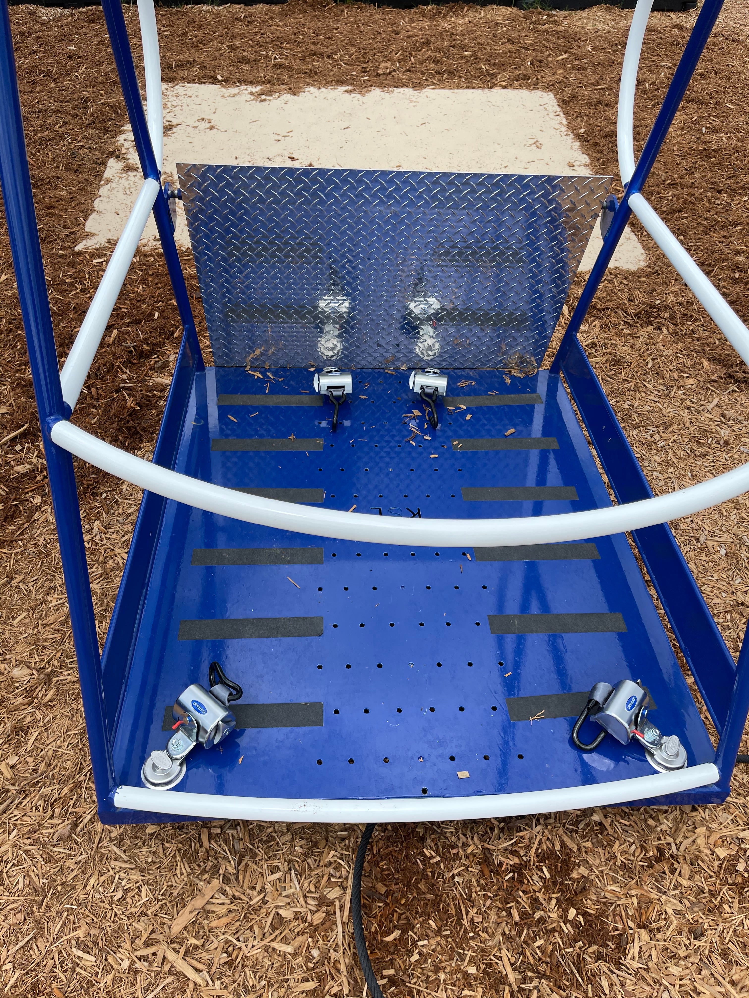 Blue and white ADA accessible swing platform with metal backrest, safety bars, and four wheel locks on playground mulch