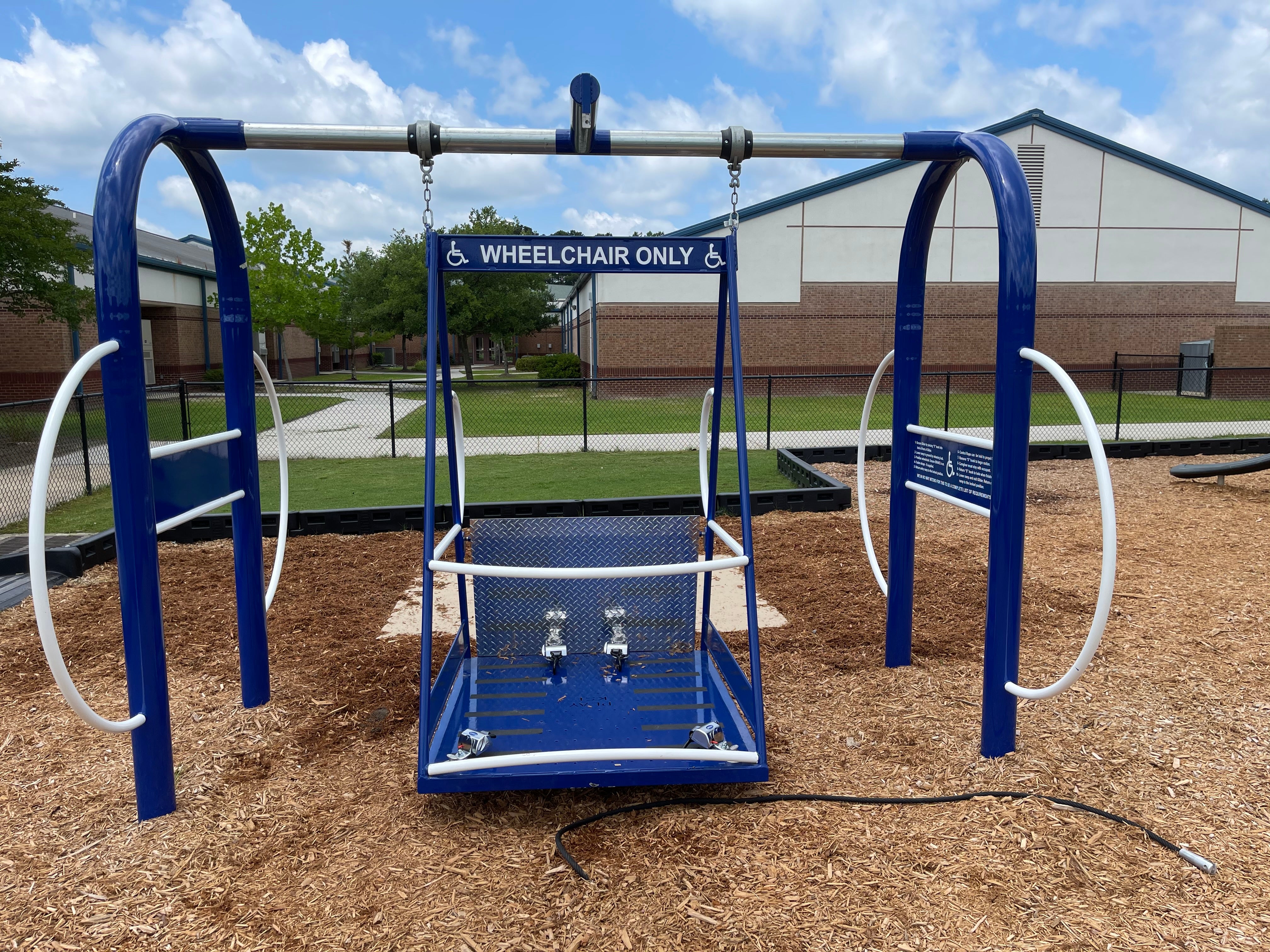Blue wheelchair-accessible playground swing with safety bars, wheelchair symbols, and wood chip ground covering