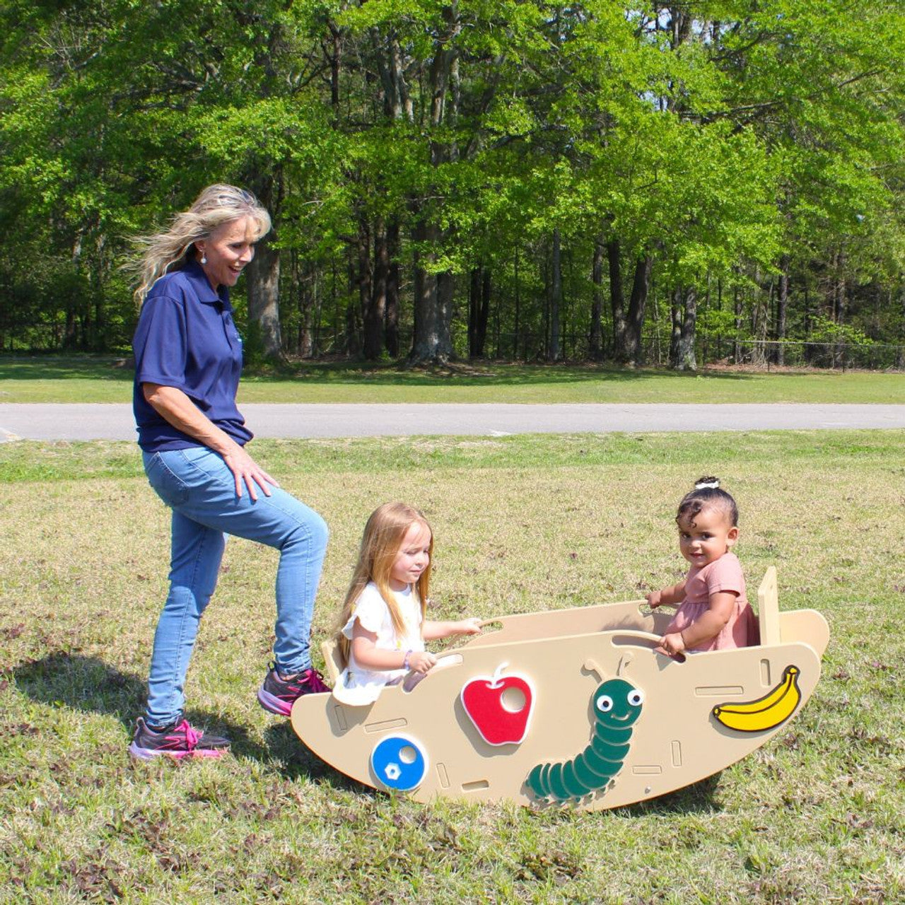 Outdoor playground seesaw with caterpillar design, hand holds, footrests, and two toddlers seated, adult assisting.