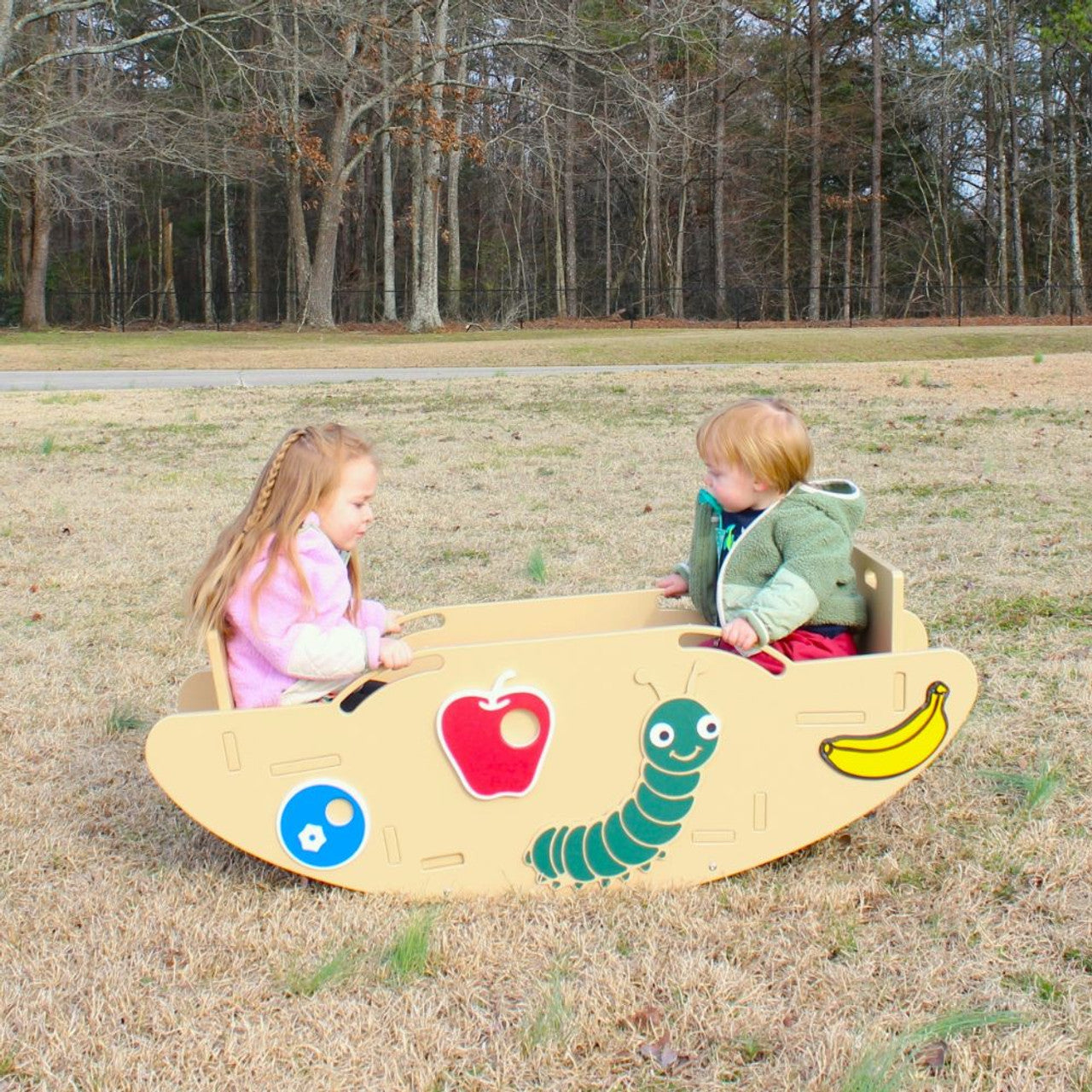 Two toddlers seated on a beige caterpillar-themed seesaw with handholds and footrests on grassy playground