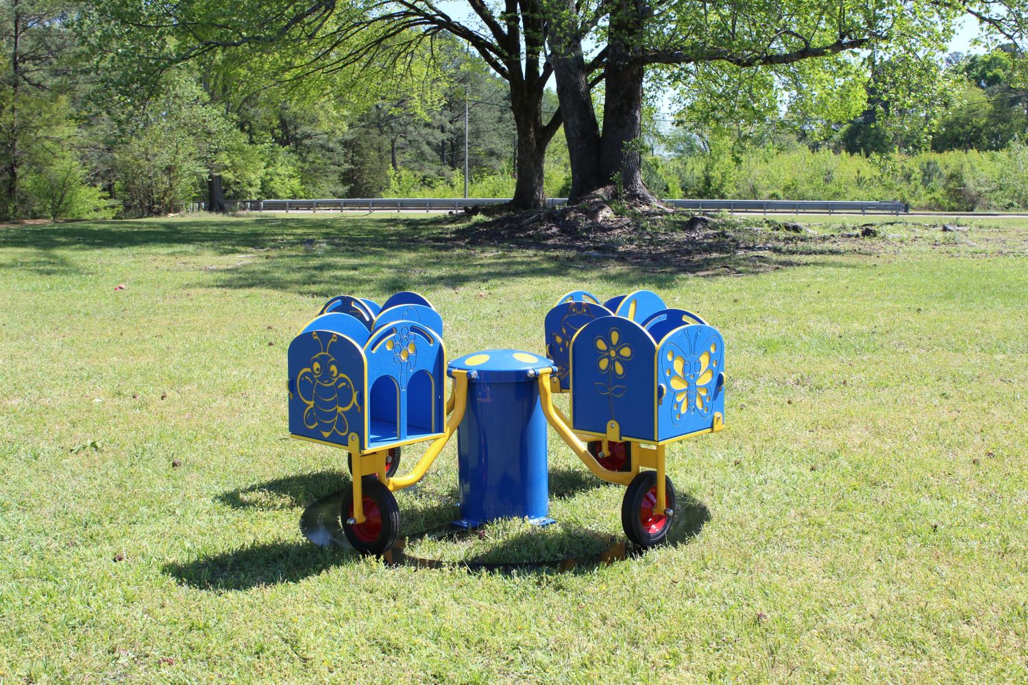 Four-seat blue and yellow merry-go-round with insect and flower designs on seats, set on grassy playground under trees