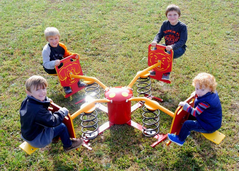 Four children seated on a red and yellow four-seat spring rider with colorful animal face designs on each seat outdoors on grass