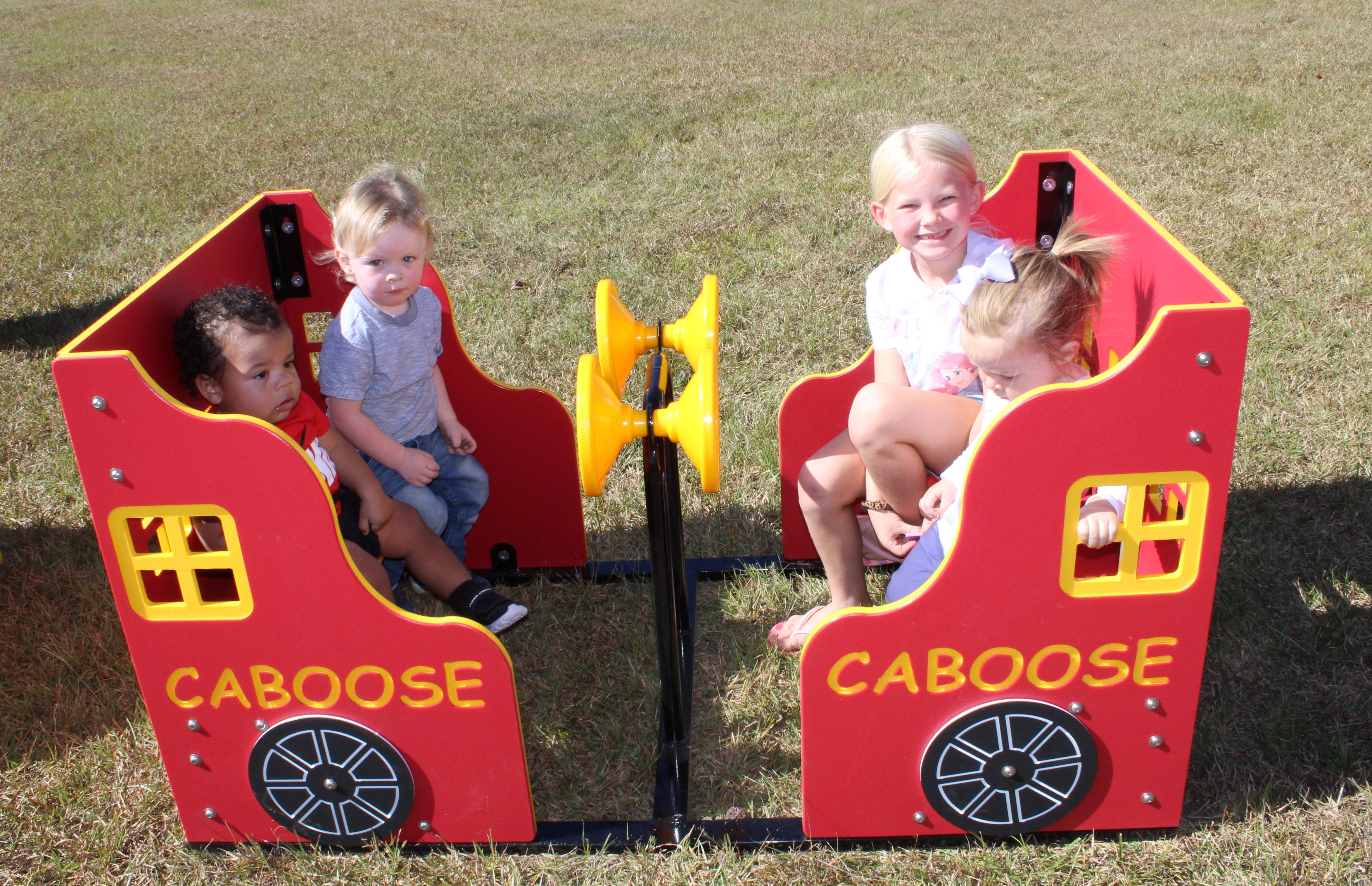 Red and yellow playground caboose with interactive wheels and four children seated on grass