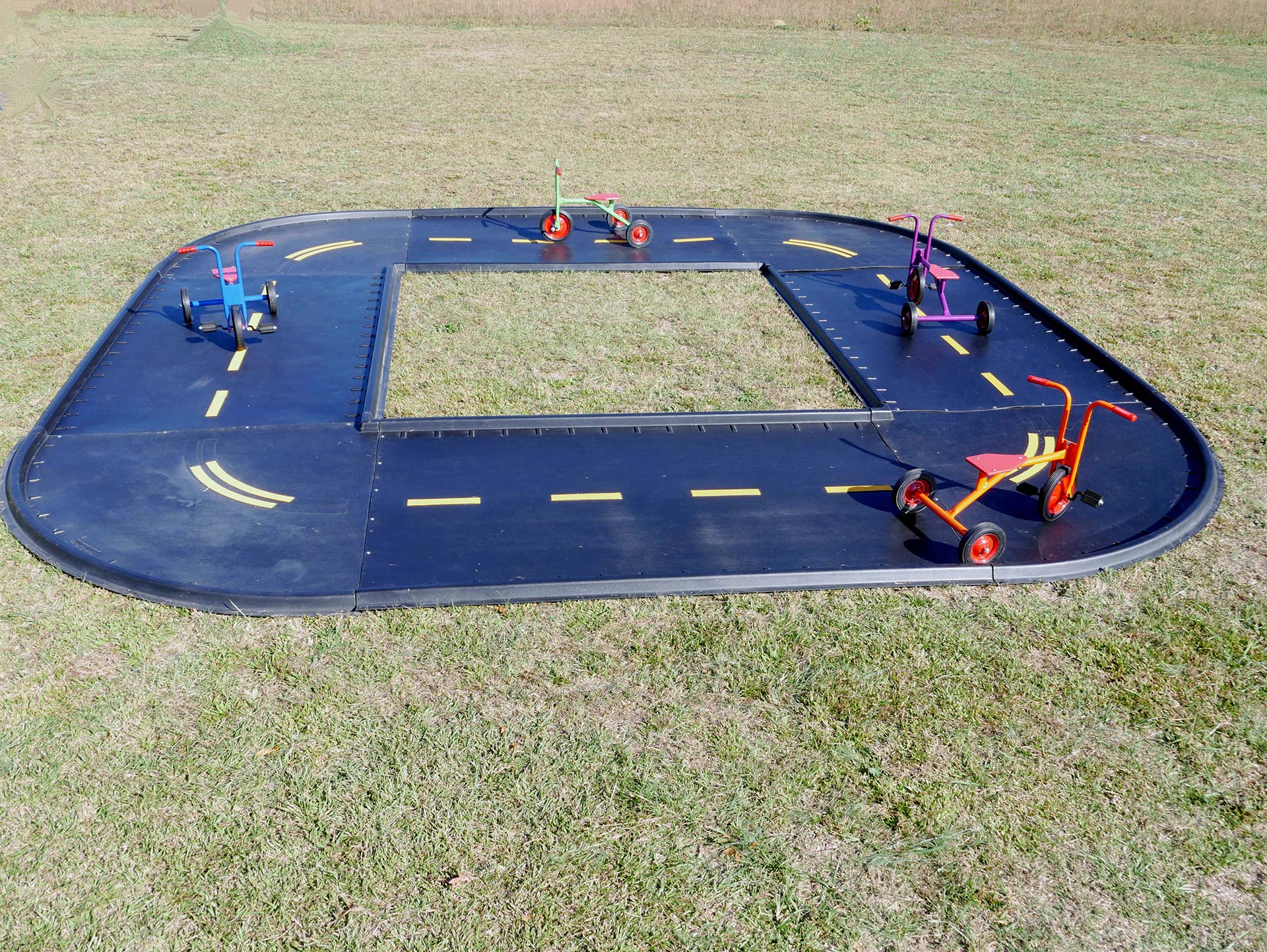 Outdoor trike path with black modular track, yellow road markings, and four colorful tricycles on grass