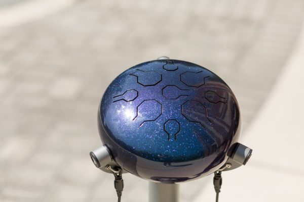 Glossy dark blue steel tongue drum with geometric cutouts mounted on a metal stand outdoors, designed for vibrant outdoor musical play