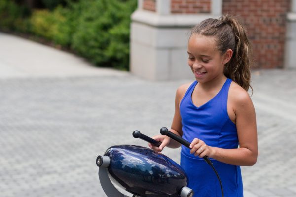 Child playing a glossy blue outdoor percussion instrument with mallets, surrounded by brick and greenery in a park setting