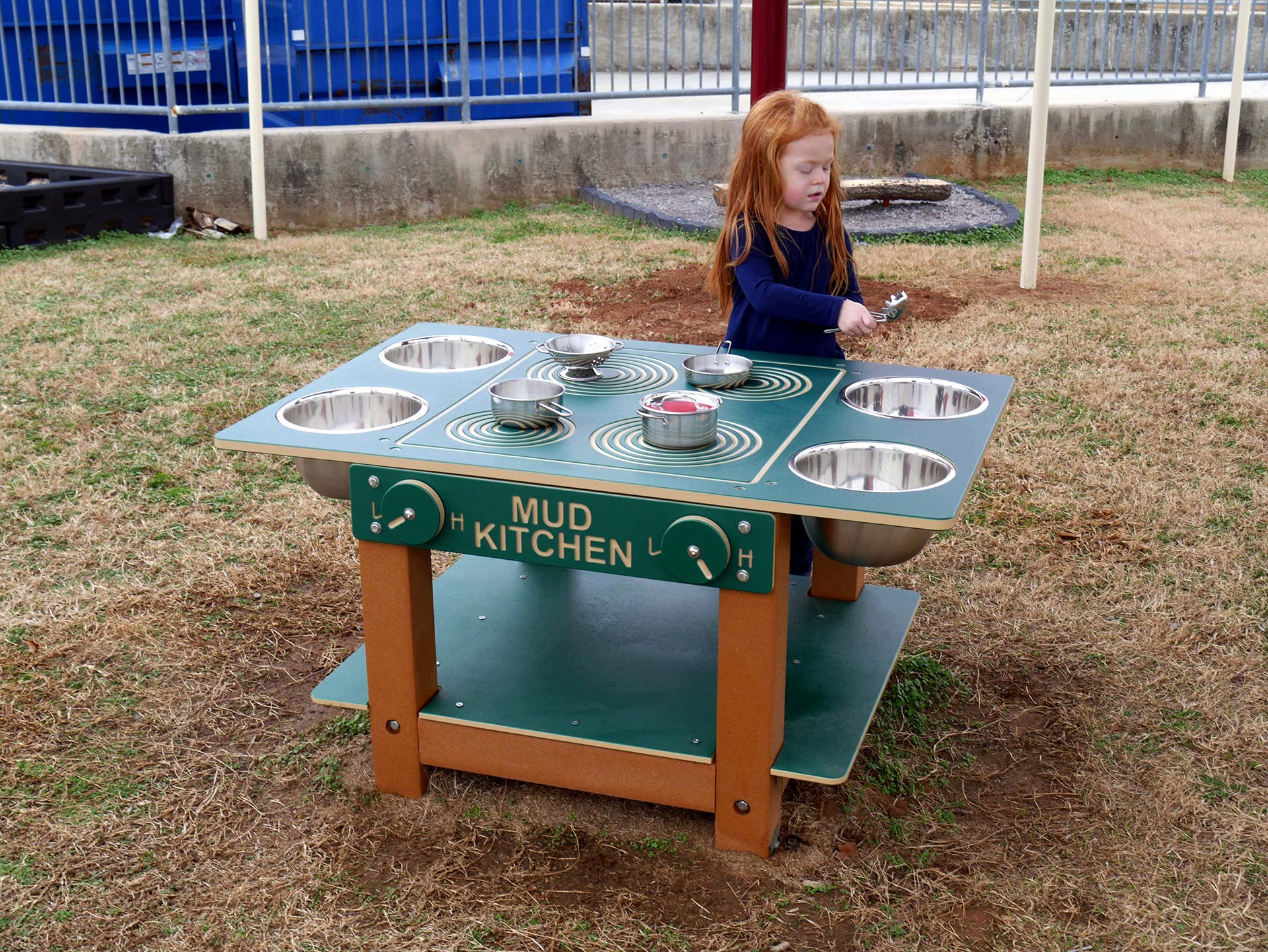 Outdoor children's mud kitchen with metal bowls, knobs, cooking utensils, and a child engaged in sensory play.