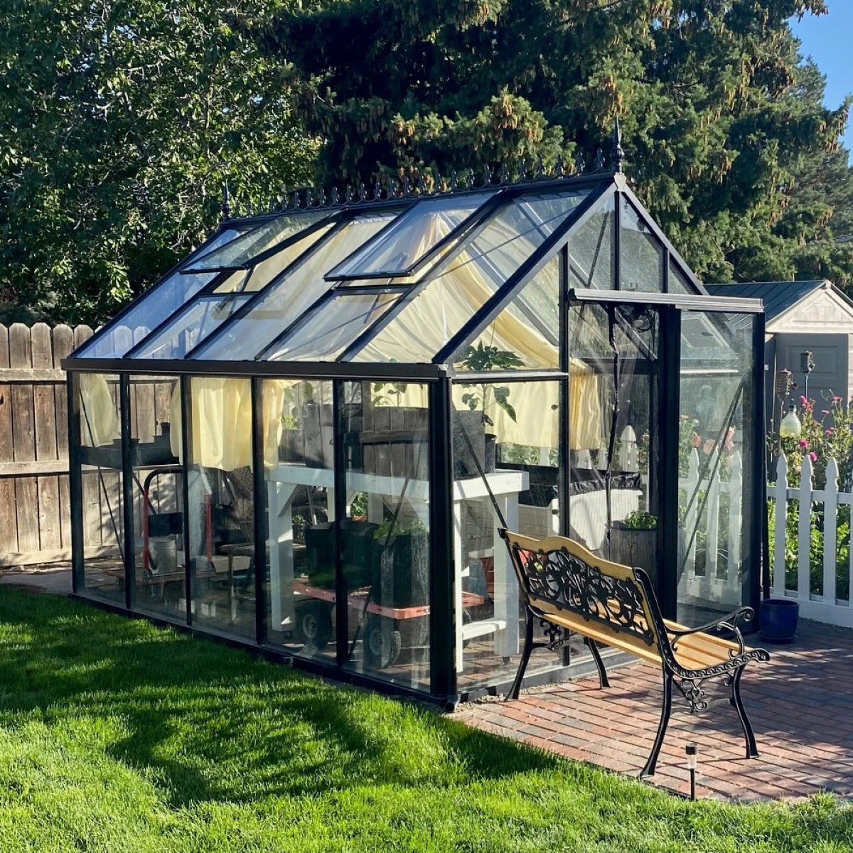 Black aluminum framed greenhouse with clear tempered glass panels, automatic roof windows, and a decorative bench on a brick patio.