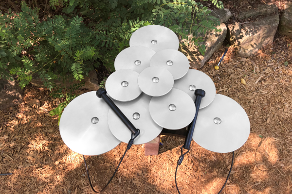 Layered silver anodized aluminum outdoor cymbals with two black mallets on wood chip ground, designed for interactive park play