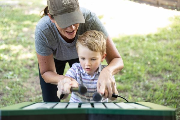Child and adult at green outdoor metallophone with nine anodized aluminum notes, set on grass in a park environment