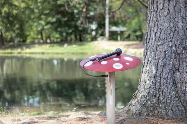 Red outdoor musical mushroom with white spots and mallet near tree and pond in natural garden setting