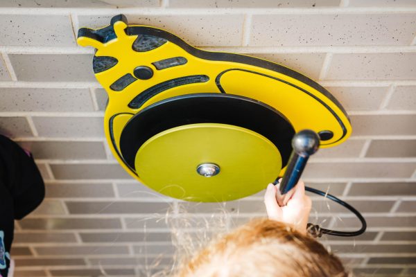 Overhead view of child playing yellow bee-shaped outdoor musical instrument with mallet on brick surface
