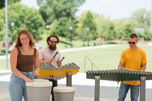 Three people playing outdoor Renaissance Ensemble musical instruments including drums and metallophones in a park setting